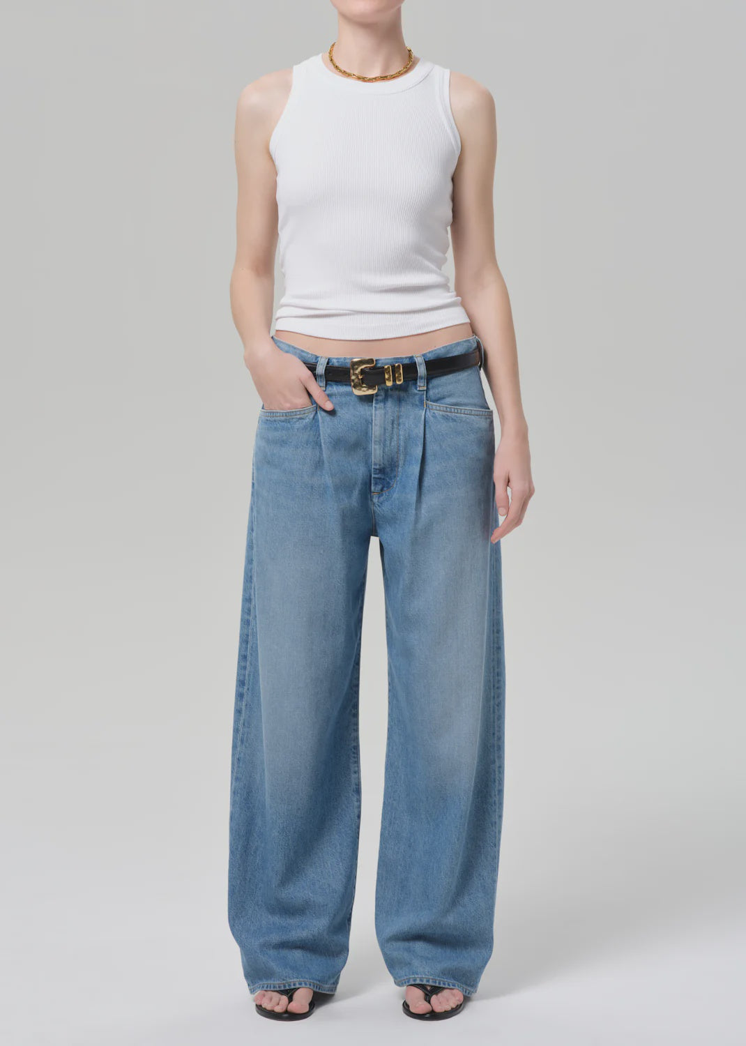 A person wearing a sleeveless white ribbed tank, black open-toe sandals, and the velha trouser in soiree by Citizens Of Humanity/AGOLDE with a black belt stands against a plain light gray background.