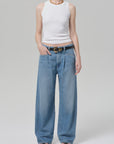 A person wearing a sleeveless white ribbed tank, black open-toe sandals, and the velha trouser in soiree by Citizens Of Humanity/AGOLDE with a black belt stands against a plain light gray background.