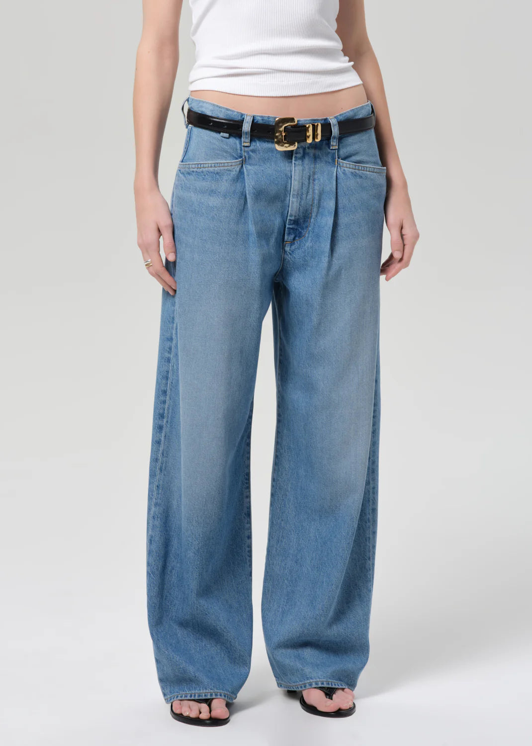 A person in a white ribbed tank top, the velha trouser in soiree by Citizens Of Humanity/AGOLDE, a black belt with a gold buckle, and black open-toe sandals stands against a plain light background.