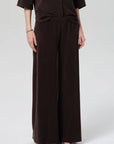 A person wears Citizens Of Humanity/AGOLDE’s nora pull on pant—a loose, wide-leg dark brown style—paired with a matching top. The relaxed lower half and part of the arms appear against a light gray background.