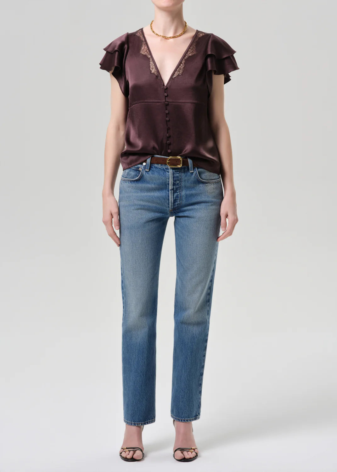 A person wears a short-sleeve, deep v-neck dark satin blouse with lace trim, Citizens Of Humanity Savannah Cigarette Jean in Patina, black heeled sandals, and a gold necklace, standing against a plain light gray background.