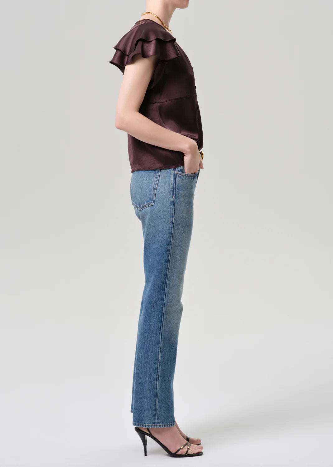 A person stands in profile wearing a short-sleeve dark ruffled top, black heeled sandals, and Citizens Of Humanity/AGOLDE’s Savannah Cigarette Jean in Patina against a plain light background.