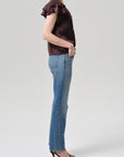 A person stands in profile wearing a short-sleeve dark ruffled top, black heeled sandals, and Citizens Of Humanity/AGOLDE’s Savannah Cigarette Jean in Patina against a plain light background.