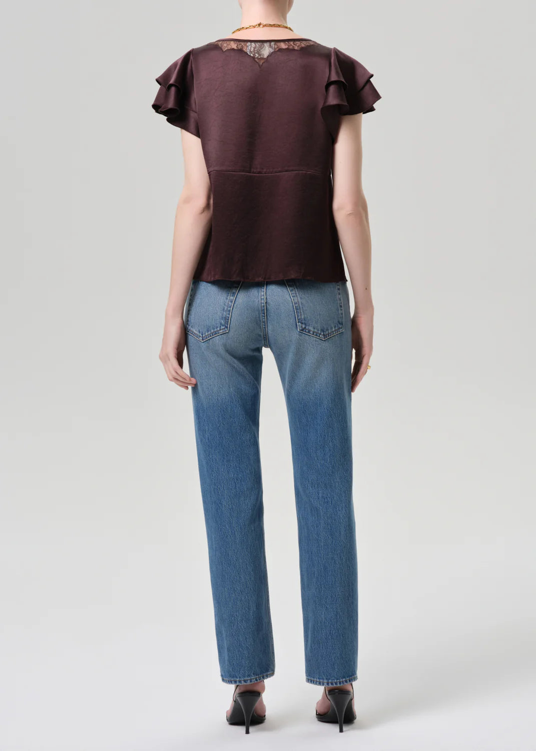 A person stands facing away in the Savannah Cigarette Jean in Patina by Citizens Of Humanity/AGOLDE, paired with a short-sleeved dark brown ruffled blouse and black heels against a plain light gray background.