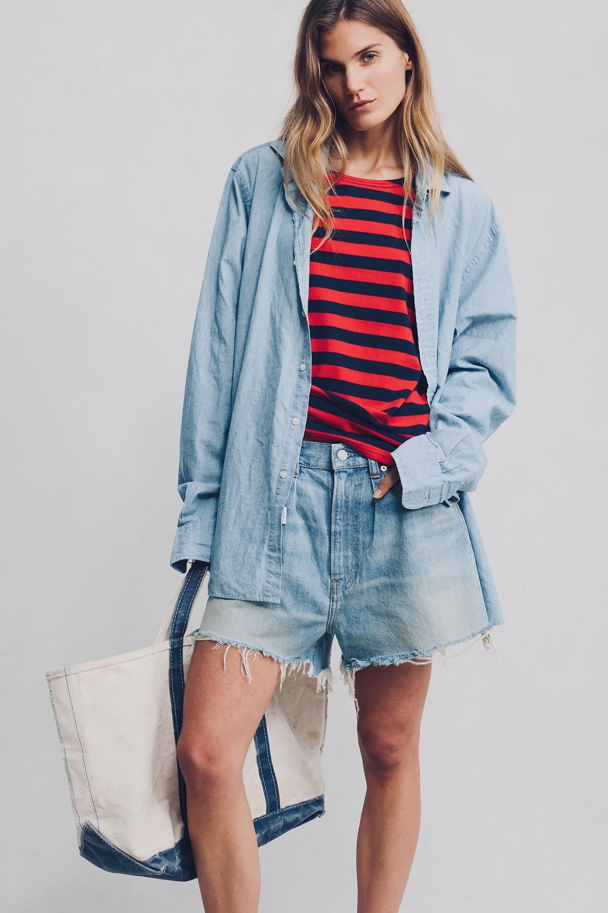 A woman in a light blue washed Maire Blue oversized button-down shirt, BROOKE SHORT MAIRE BLUE by Denimist with a raw hem, and holding a large white canvas tote bag with blue trim stands against a plain background.