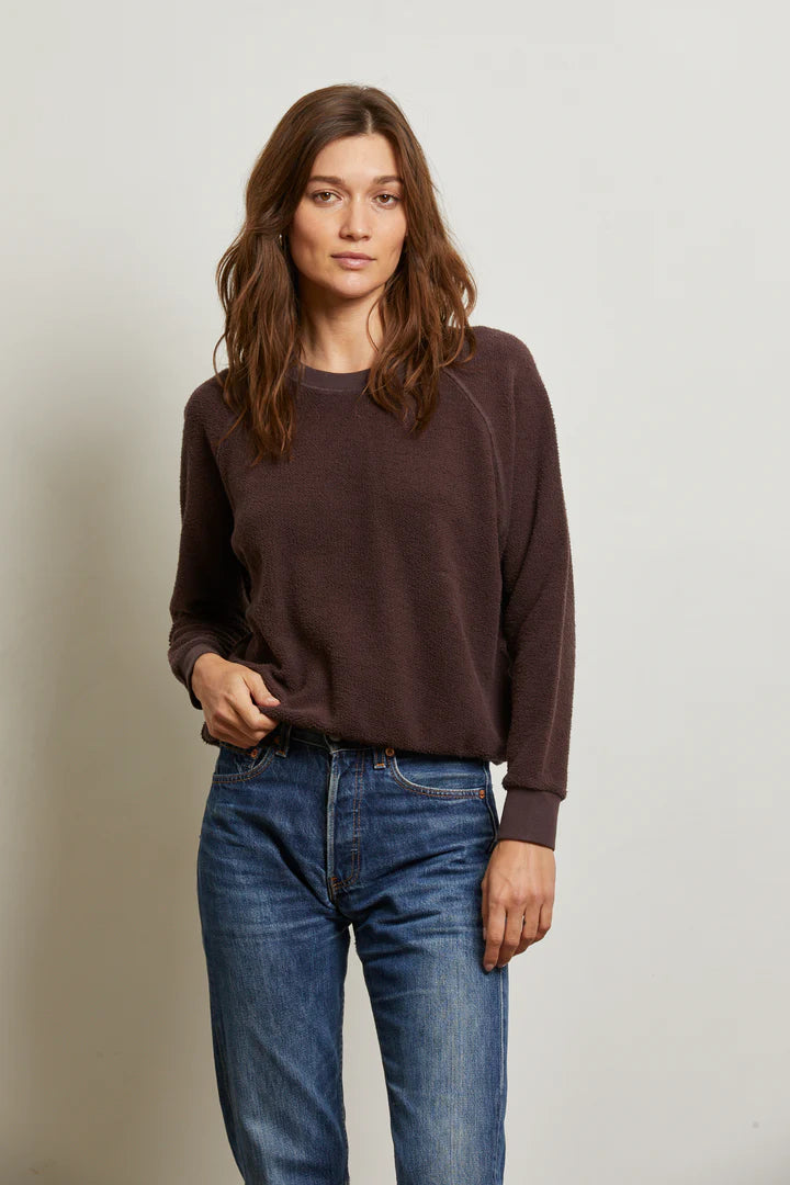 A woman with long brown hair wears the Perfectwhitetee Ziggy Reverse LS Sweatshirt in dark brown with bracelet-length sleeves and blue jeans, standing against a plain light background, looking at the camera neutrally.