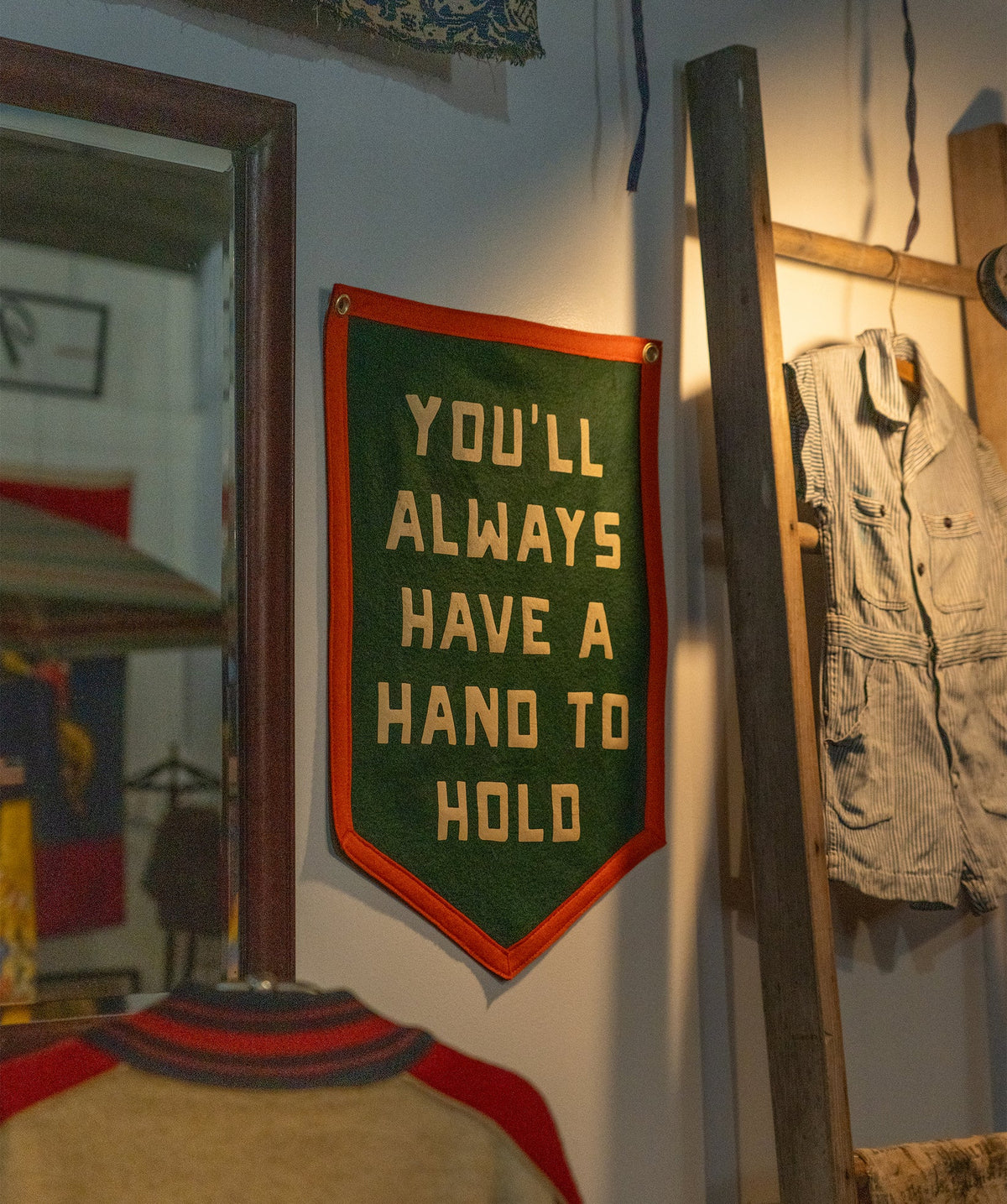 The Oxford Pennant "You'll Always Have a Hand to Hold" camp flag in green and orange adds a cozy touch to your space, displayed near a wooden ladder, striped shirt, and mirror in a warmly lit room.