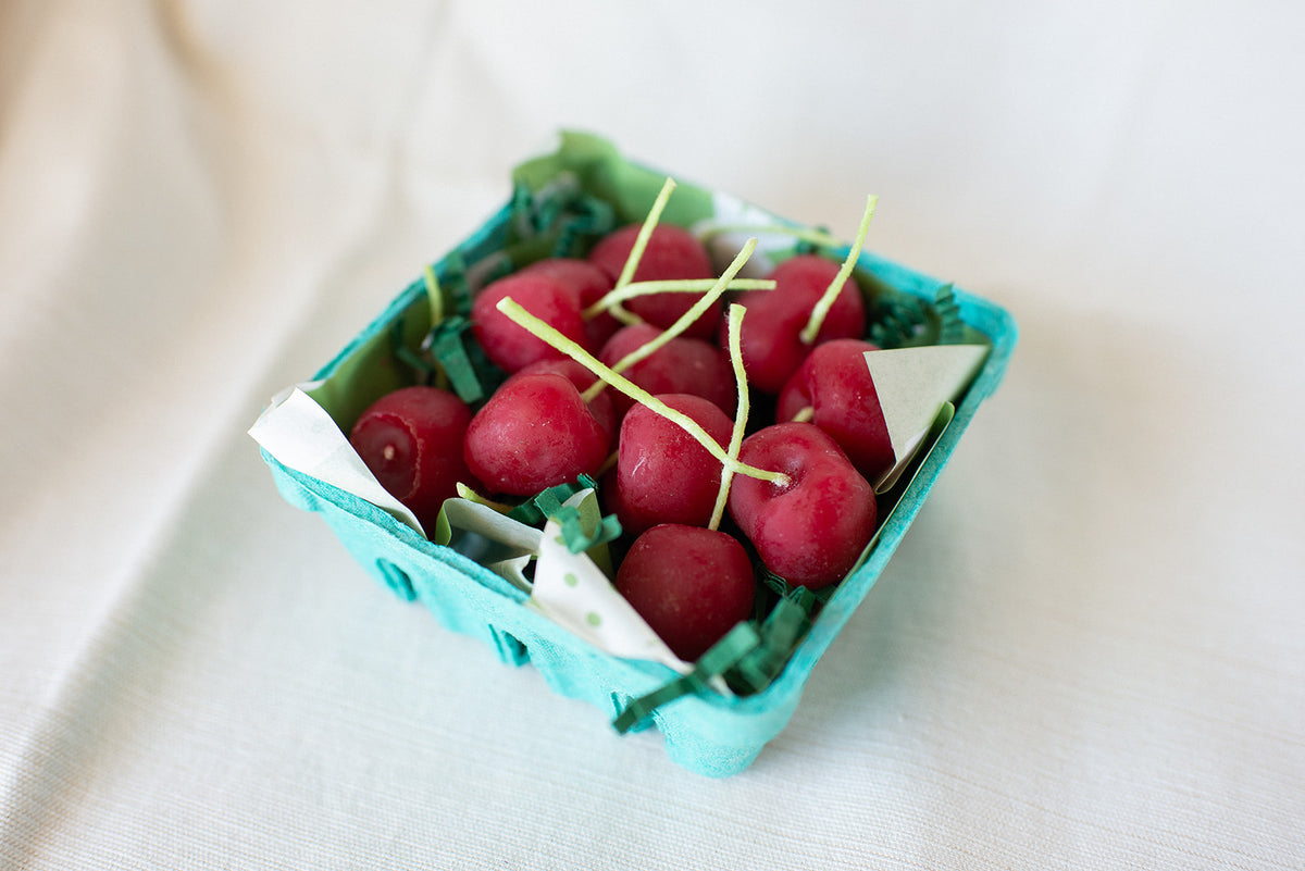 A turquoise carton from Faire holds Cherry Beeswax Candles—handmade, cherry-shaped with green stems—resting on white fabric and cushioned by green paper shreds for a playful, charming display.