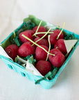 A turquoise carton from Faire holds Cherry Beeswax Candles—handmade, cherry-shaped with green stems—resting on white fabric and cushioned by green paper shreds for a playful, charming display.