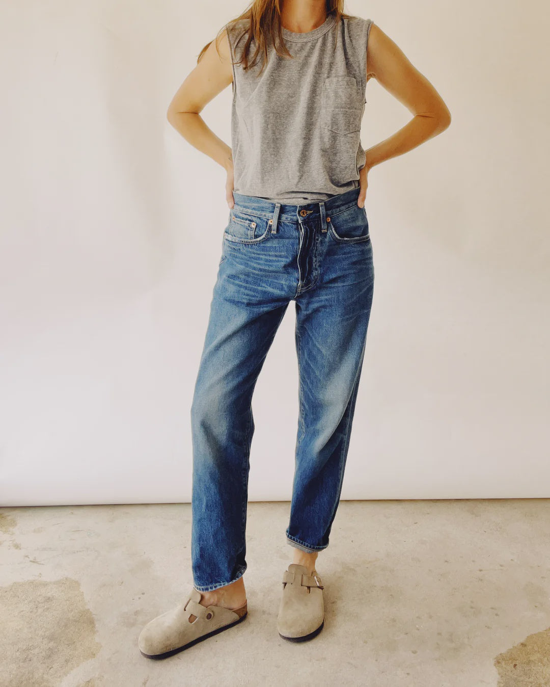 A person confidently stands with hands on hips in Hey Gang's "The Loose Crop," a gray sleeveless top, and 100% cotton mid-rise relaxed jeans, their light brown clogs grounding them on the concrete floor. The neutral background highlights the stylish ensemble.