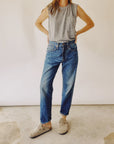 A person confidently stands with hands on hips in Hey Gang's "The Loose Crop," a gray sleeveless top, and 100% cotton mid-rise relaxed jeans, their light brown clogs grounding them on the concrete floor. The neutral background highlights the stylish ensemble.