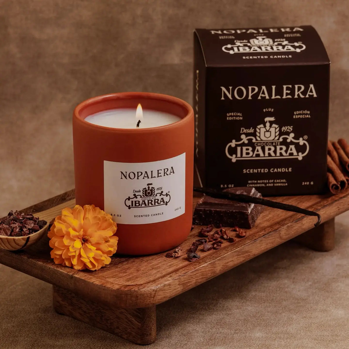 A Nopalera Chocolate Candle by Faire, in a brown glass jar, sits on a tray with marigold, cinnamon sticks, and chocolate pieces. The box highlights the “Chocolate Ibarra” scent inspired by Mexican heritage.