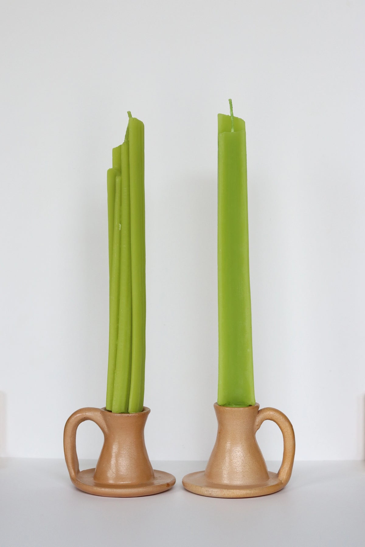 Two light brown ceramic holders cradle Faire’s handmade Celery Beeswax Candles—one green taper is bent and uneven, the other stands straight—displayed against a plain white background.
