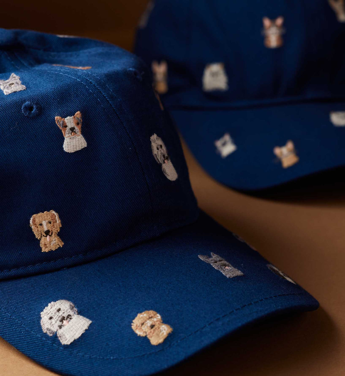 Close-up of two blue Faire Dog Days Baseball Caps with small, embroidered cartoon dog faces in different breeds and colors. Each cap features an adjustable strap and rests on a brown surface.