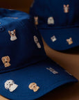 Close-up of two blue Faire Dog Days Baseball Caps with small, embroidered cartoon dog faces in different breeds and colors. Each cap features an adjustable strap and rests on a brown surface.