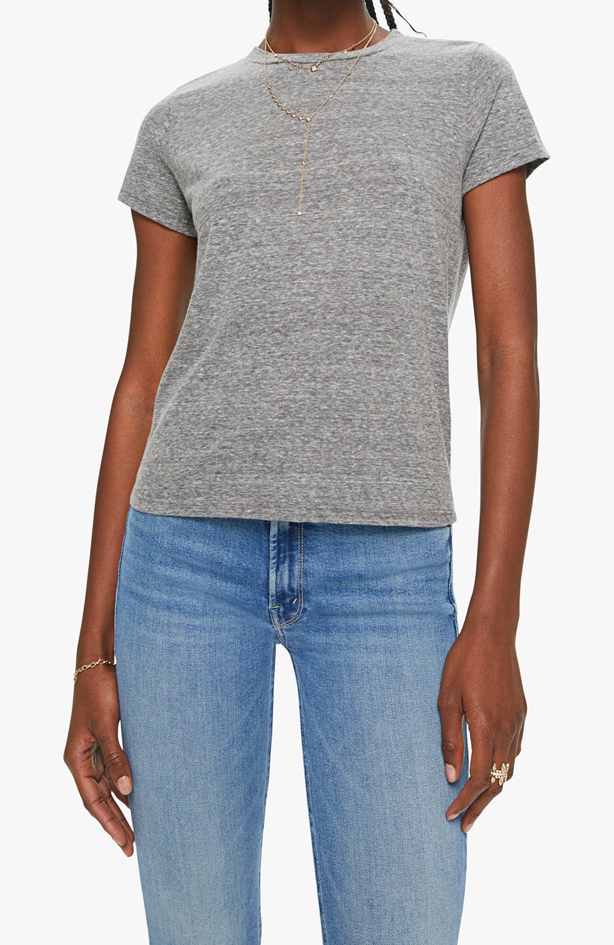 Dressed in Mother’s The Lil Goodie Goodie Tee in heather gray with a crewneck, blue jeans, gold layered necklaces, bracelet, and rings, a person stands against a plain light background for an effortless vintage style.