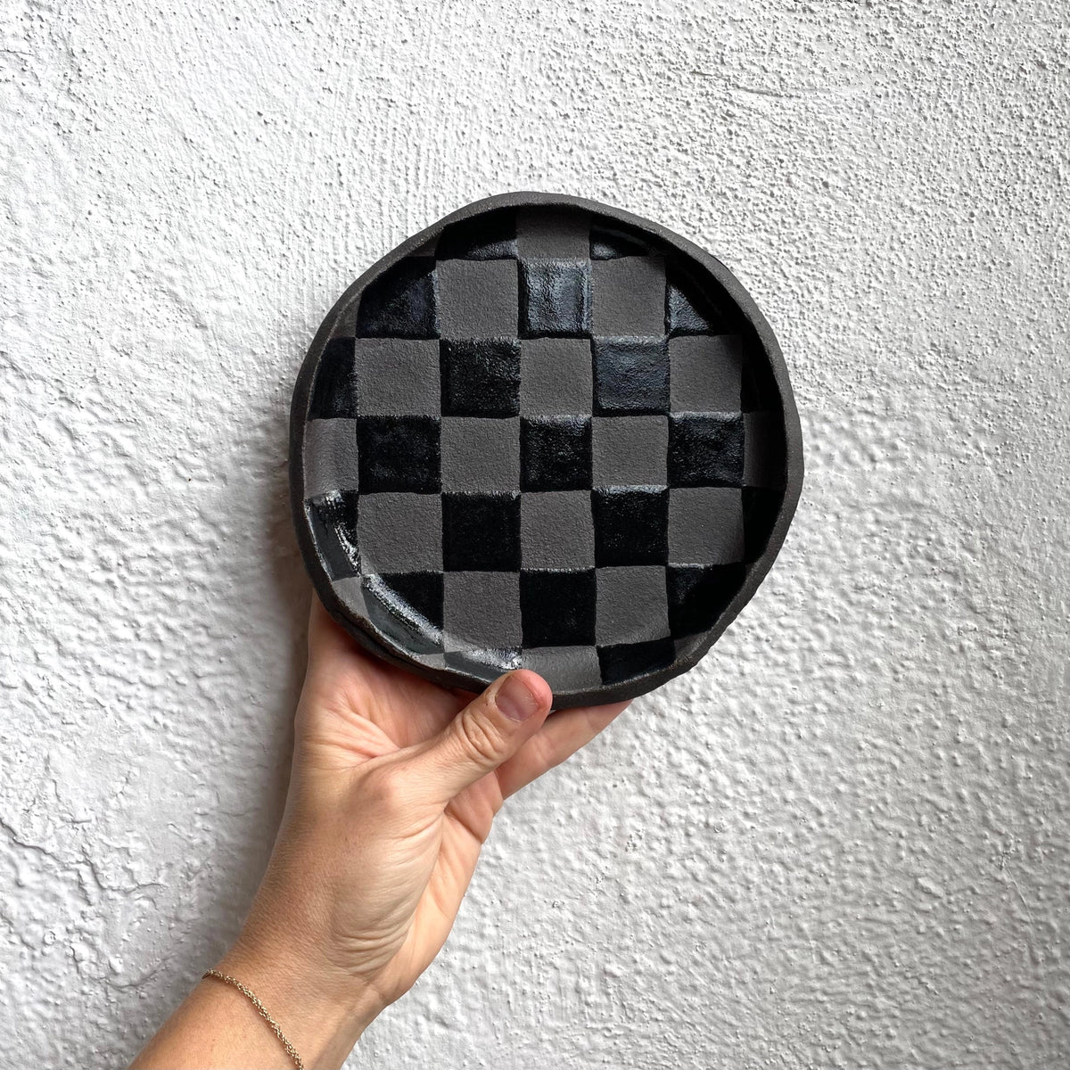 Black Check Snack Plate by Faire with a hand holding it against a textured white wall.