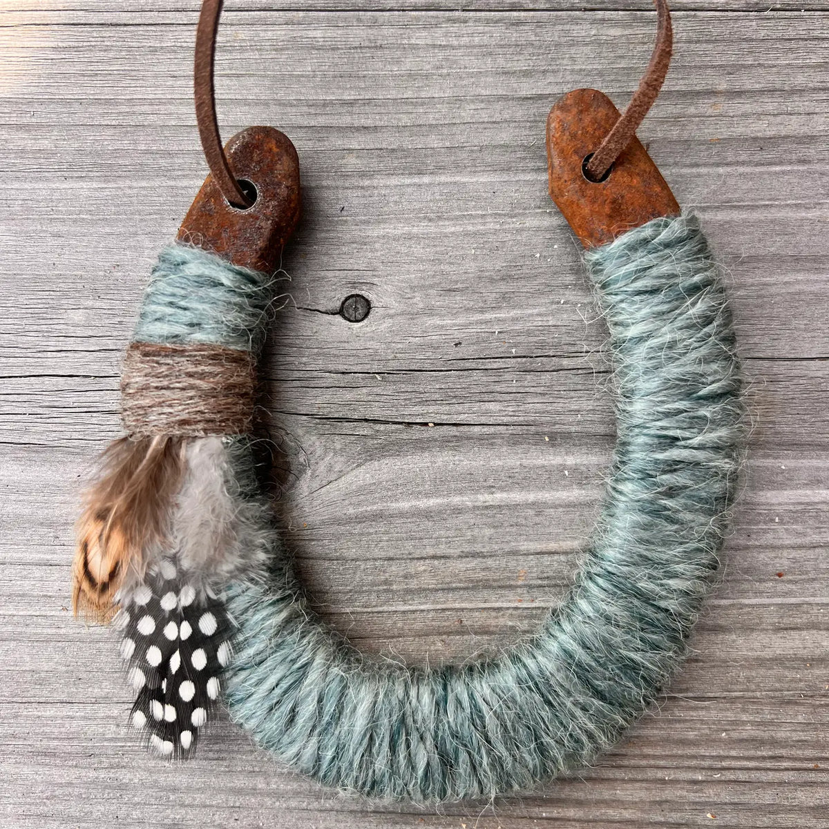 The Lucky Horseshoe by Faire features an upcycled horseshoe wrapped in blue-grey twine, brown string, and feathers, hanging from a leather cord—a unique housewarming or personalized gift.