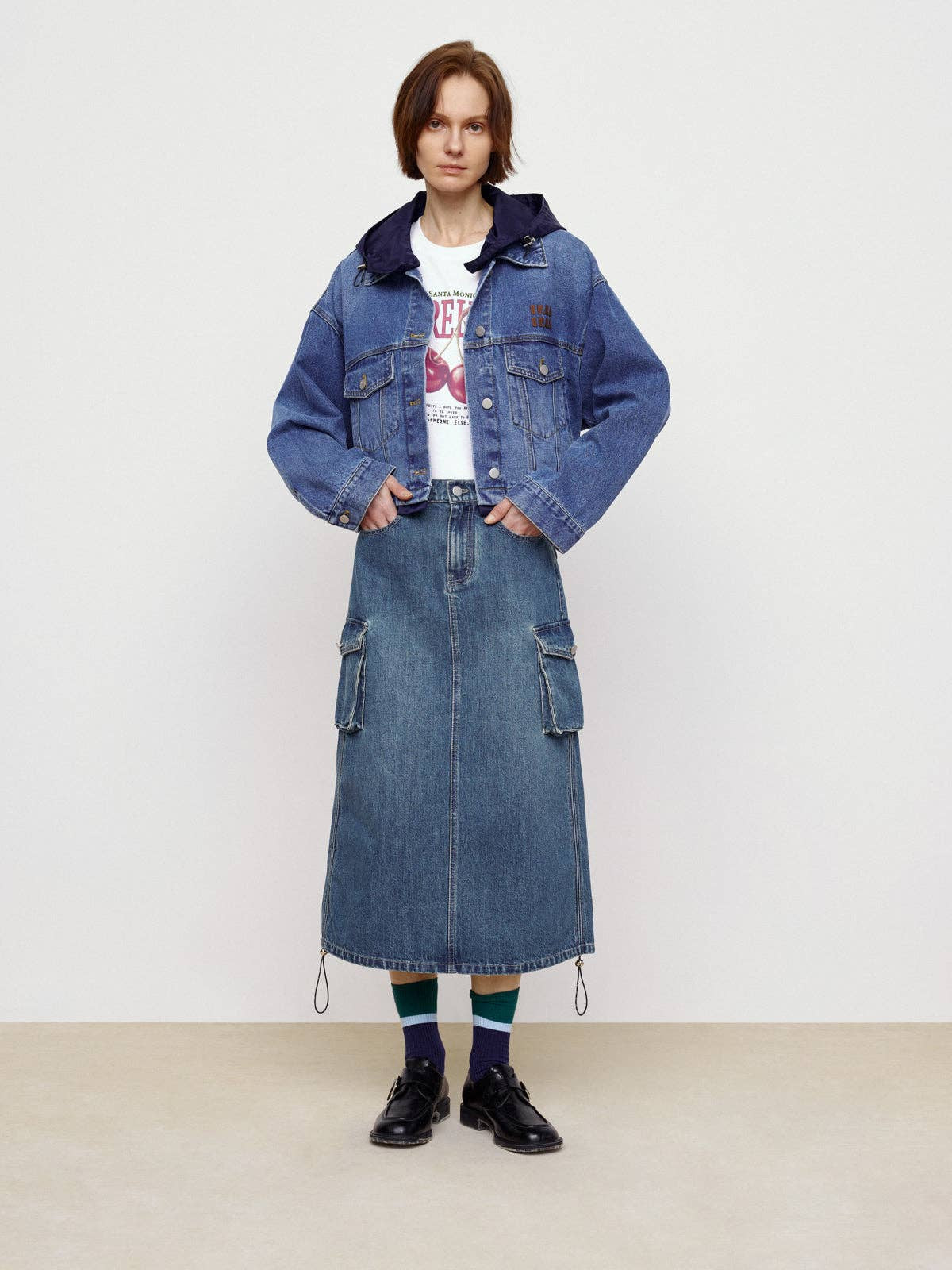 A person stands against a plain background, showcasing street style in a blue denim jacket, white graphic tee, Faire's Pocket Denim Half Skirt, blue and green socks, and black shoes.