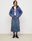 A person stands against a plain background, showcasing street style in a blue denim jacket, white graphic tee, Faire's Pocket Denim Half Skirt, blue and green socks, and black shoes.