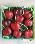 A small blue basket by Faire holds Cherry Beeswax Candles shaped like red and dark red hearts with green candy stems, arranged on green paper shreds.