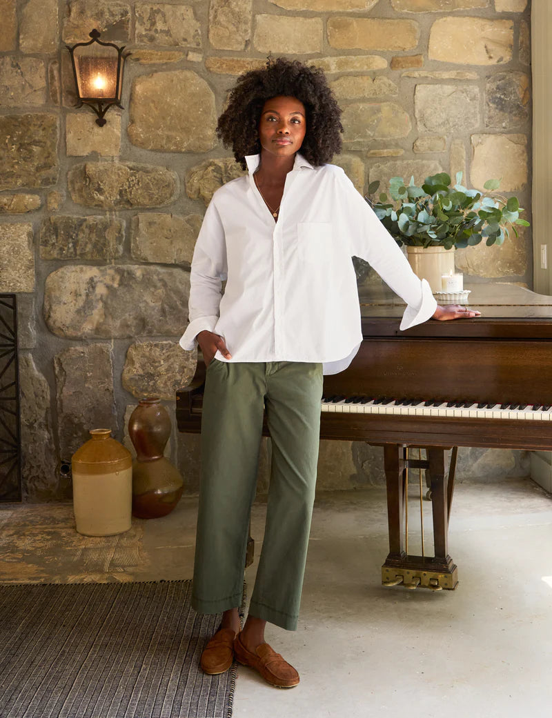 A person with curly hair, wearing a relaxed-fit, 100% cotton Oona Continuous Button-Up Shirt by Frank &amp; Eileen and green pants, stands with one hand resting on a piano in a room with stone walls. There is a plant on the piano and two large vases on the floor nearby. A lit wall sconce adds warm light to the scene.