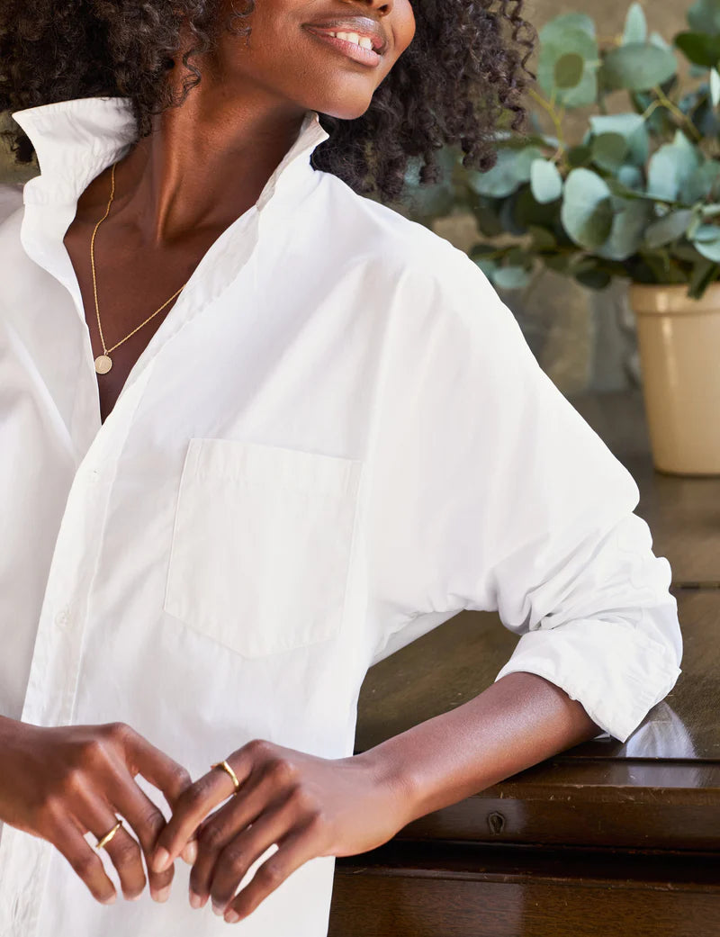 A person with curly hair is leaning against a wooden surface. They are wearing the Oona Continuous Button-Up Shirt made from 100% cotton in white, by Frank &amp; Eileen, with the collar popped up and sleeves rolled. The person also wears gold rings and a delicate gold necklace. A potted plant is blurred in the background.