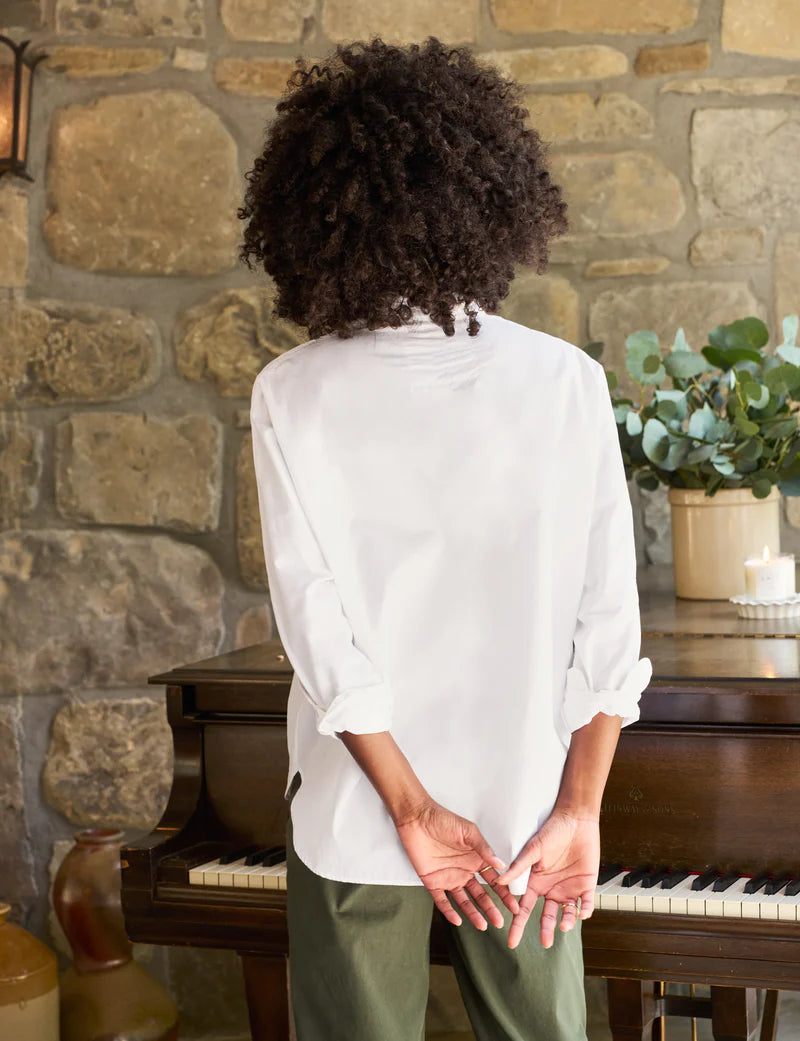 A person with curly hair is standing with their back turned, facing a stone wall and an upright piano adorned with decorative plants on top. They are wearing a Frank &amp; Eileen Oona Continuous Button-Up Shirt made from 100% cotton and green pants, with their hands clasped behind their back.
