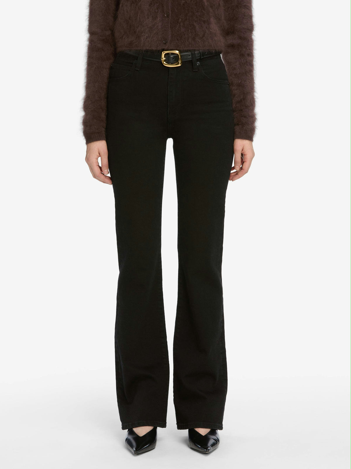 A person wears black bootcut jeans, a fuzzy dark sweater, and pointed black heels, styled with FRAME's The Organic Buckle Belt featuring a gold buckle, all set against a white background.