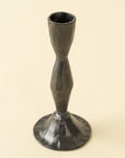 The Zora Forged Candlestick L by Indaba features a dark metallic finish, a textured surface, narrow midsection, and wide flared base, standing upright against a plain light background.