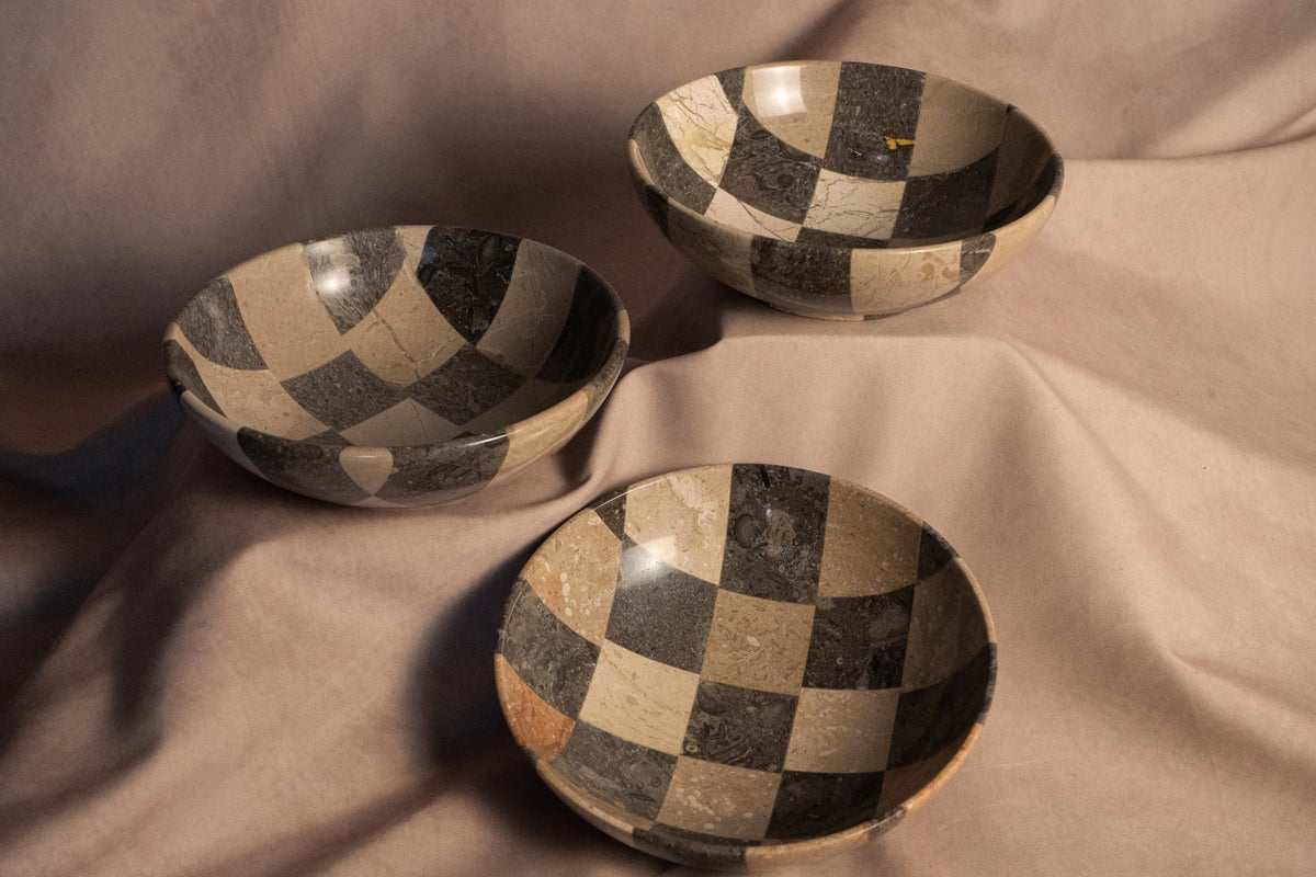 Hand-finished checkered travertine bowl by The Faire on beige fabric, classic accent for living or dining spaces.