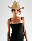 A woman in the Janessa Leone Sacha Hat and a black spaghetti strap dress stands against a plain, light background, looking directly at the camera with a neutral expression.