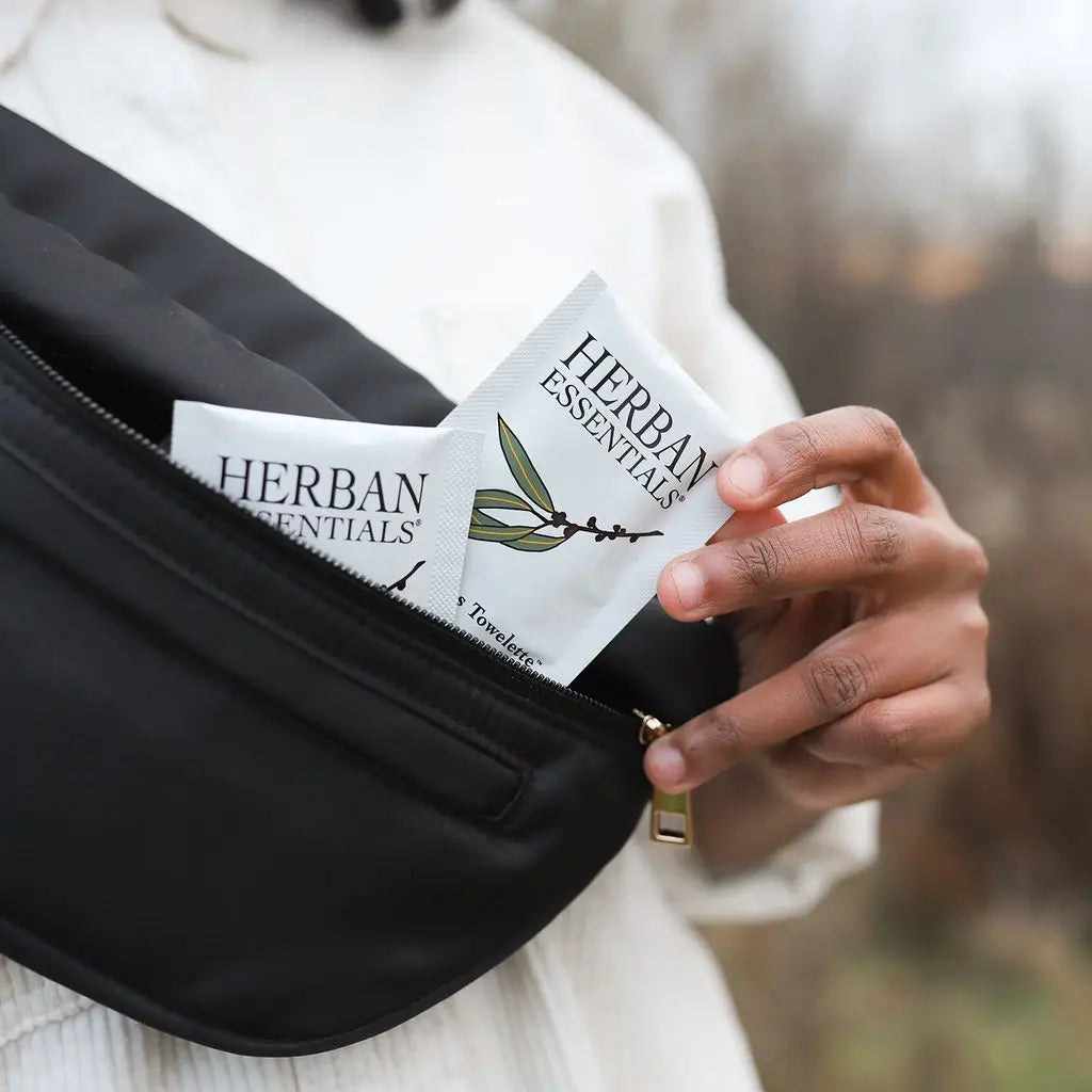 A person in a white shirt pulls a Faire Eucalyptus Essential Oil Towelettes | Portable Aromatherapy packet from a black zippered bag, with another travel refreshing wipe also visible inside.