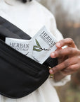 A person in a white shirt pulls a Faire Eucalyptus Essential Oil Towelettes | Portable Aromatherapy packet from a black zippered bag, with another travel refreshing wipe also visible inside.