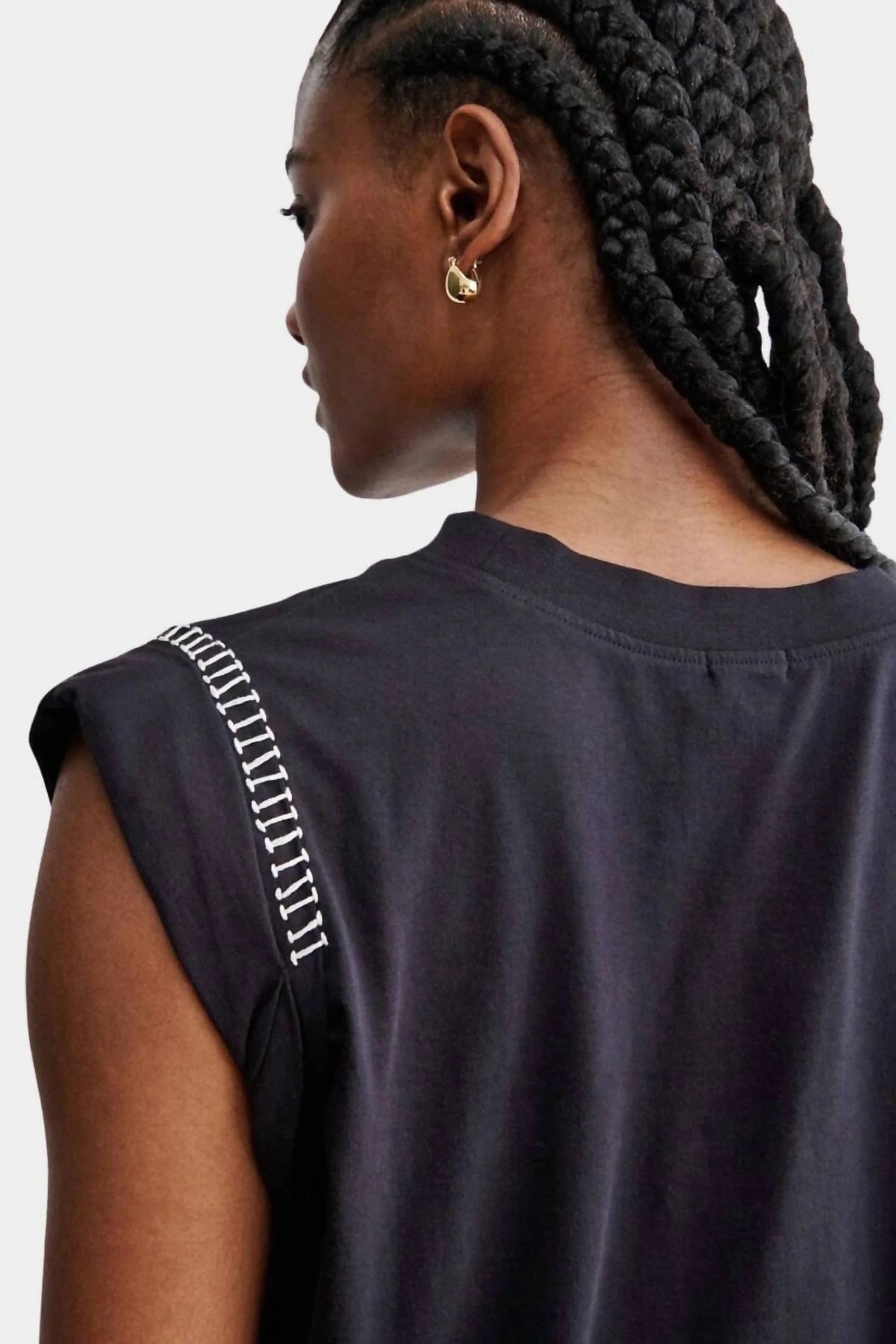 A person with braided hair is seen from the back, wearing a black Rachel Comey Miles Tee in a minimalist style with white hand crochet stitch detail on the shoulder seam and a gold hoop earring.