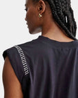 A person with braided hair is seen from the back, wearing a black Rachel Comey Miles Tee in a minimalist style with white hand crochet stitch detail on the shoulder seam and a gold hoop earring.