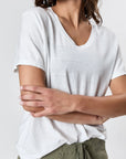 A person in the Faire Hemp V Neck t-shirt and olive green drawstring pants stands with arms crossed, facing slightly to the side against a plain white background. Their face is partially visible.