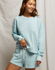 A person with long hair smiles while wearing the Layla Terry Short from Perfectwhitetee, paired with a light blue sweatshirt. Crafted from soft French terry cotton, they stand confidently before a light fabric backdrop, hand resting on their sweatshirt hem, showcasing effortless comfort.