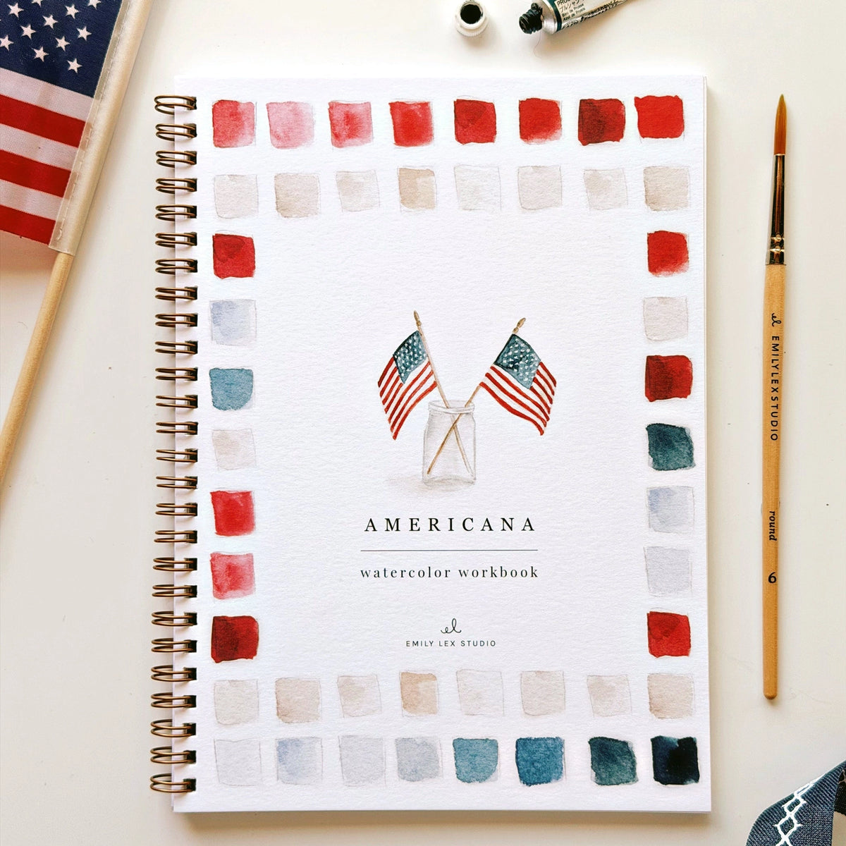 The Faire Watercolor Workbook, a spiral-bound beginner guide titled "Americana," features two small American flags in a glass jar on the cover with red, blue, and gray watercolor swatches, shown on a table with paint supplies.