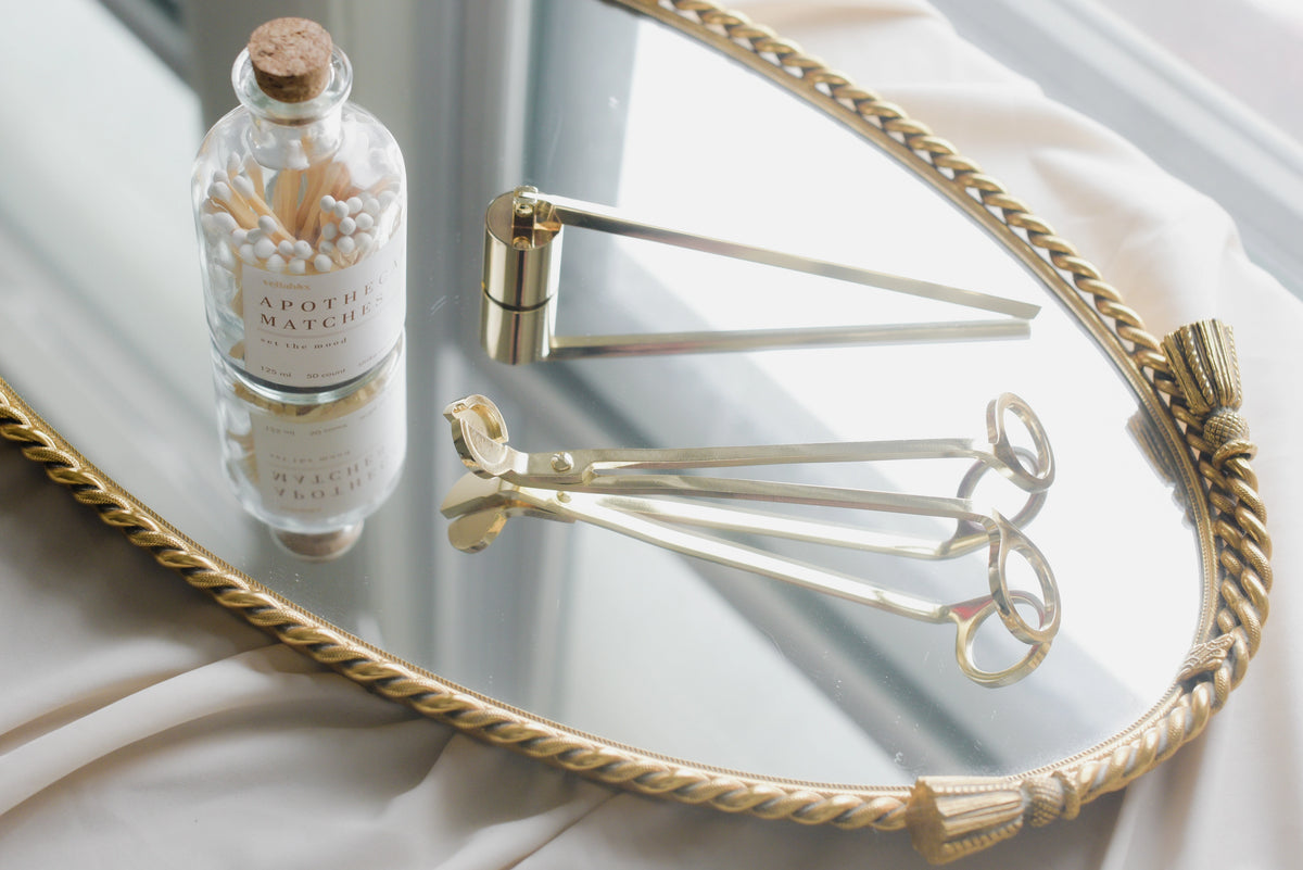 A Faire Candle Snuffer rests with two gold wick trimmers on an ornate oval mirror tray near a sunlit window, showcasing elegant and functional candle accessories for stylish home decor.