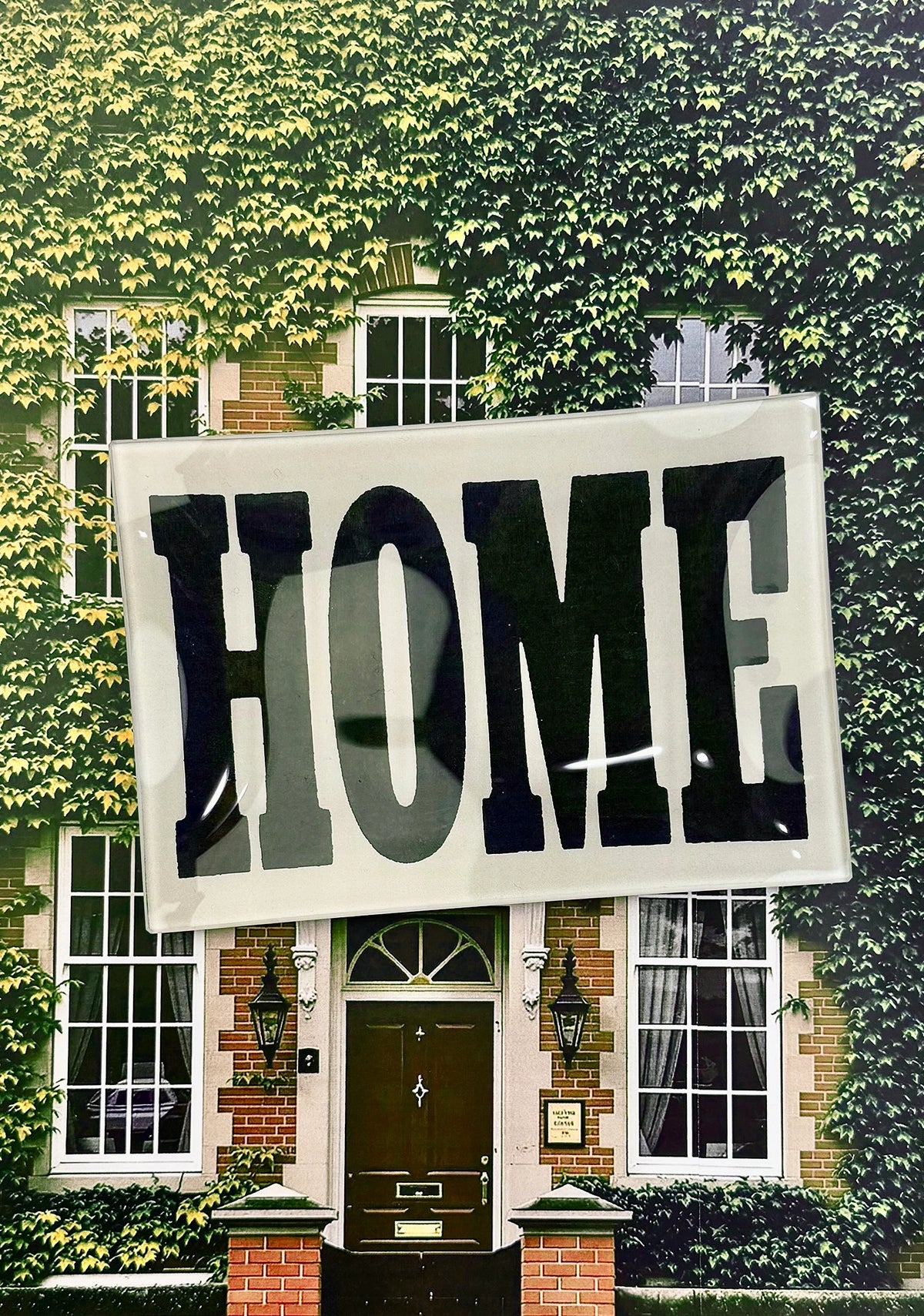 The Faire Home Block Decoupage Glass Tray, featuring vintage "HOME" typography, is displayed in front of a green ivy-clad house with brick walls, large windows, and a dark brown door.