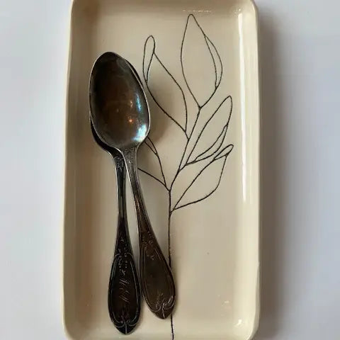 Two hand-etched silver spoons rest on Faire’s Floral Small Tray, a rectangular cream-colored piece featuring a minimalist black line drawing of a leafy branch. The spoons’ handles align perfectly with the base of the drawn branch.
