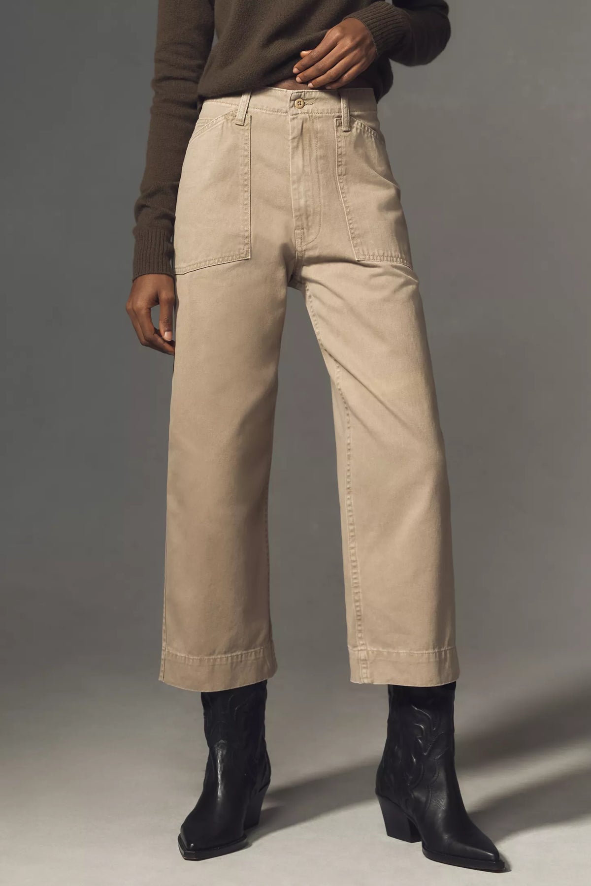 A person wears the Denimist Georgia Work Pant—beige, high-waisted, wide-leg pants with front patch pockets—with a brown long-sleeve top and black ankle boots, standing against a plain gray background.