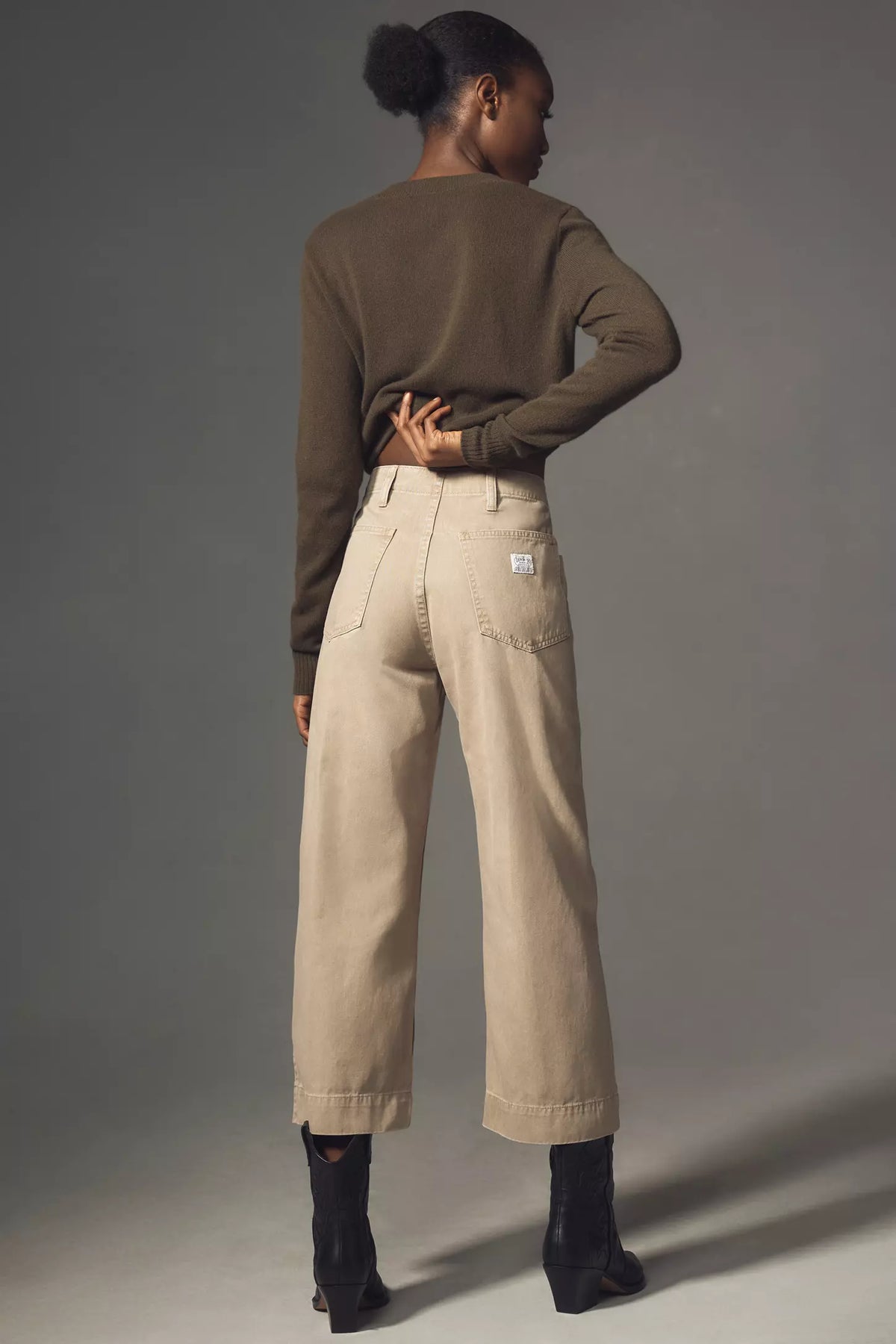 A person stands against a plain gray background, wearing Denimist's Georgia Work Pant—a high-waisted, wide-leg style—paired with a brown sweater and black ankle boots, with one hand on their hip.