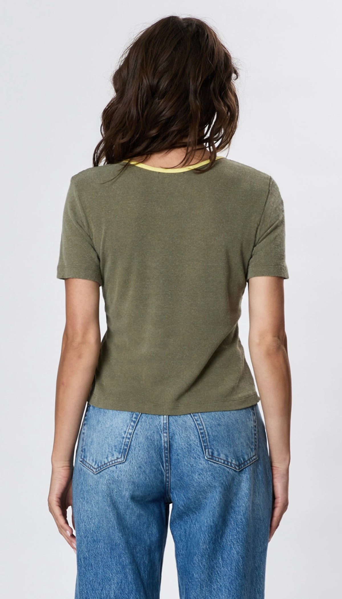 A woman with wavy brown hair is shown from the back, wearing Faire’s Stardust Hemp Top—an olive green short-sleeve tee with a yellow neckline—plus high-waisted blue jeans, radiating retro charm against a light background.
