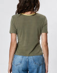 A woman with wavy brown hair is shown from the back, wearing Faire’s Stardust Hemp Top—an olive green short-sleeve tee with a yellow neckline—plus high-waisted blue jeans, radiating retro charm against a light background.