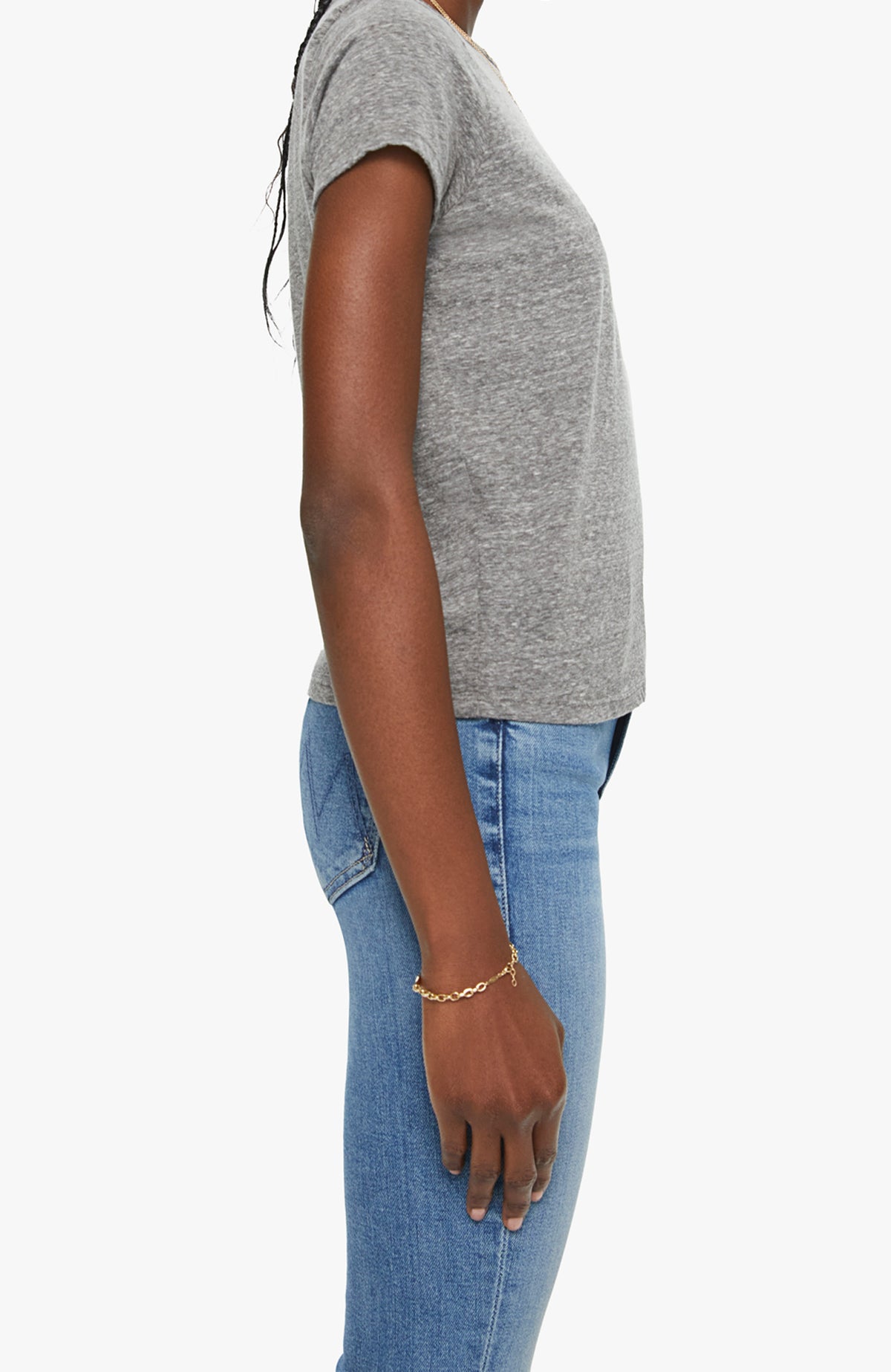 A person stands in profile against a plain background, wearing Mother’s The Lil Goodie Goodie Tee in grey. Their right arm hangs relaxed by their side, embodying the timeless ease of this essential short-sleeve crewneck.