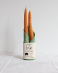Two orange Carrot Beeswax Candles by Faire, made from beeswax, are wrapped in green paper and stand upright on a white textured surface against a plain white background.