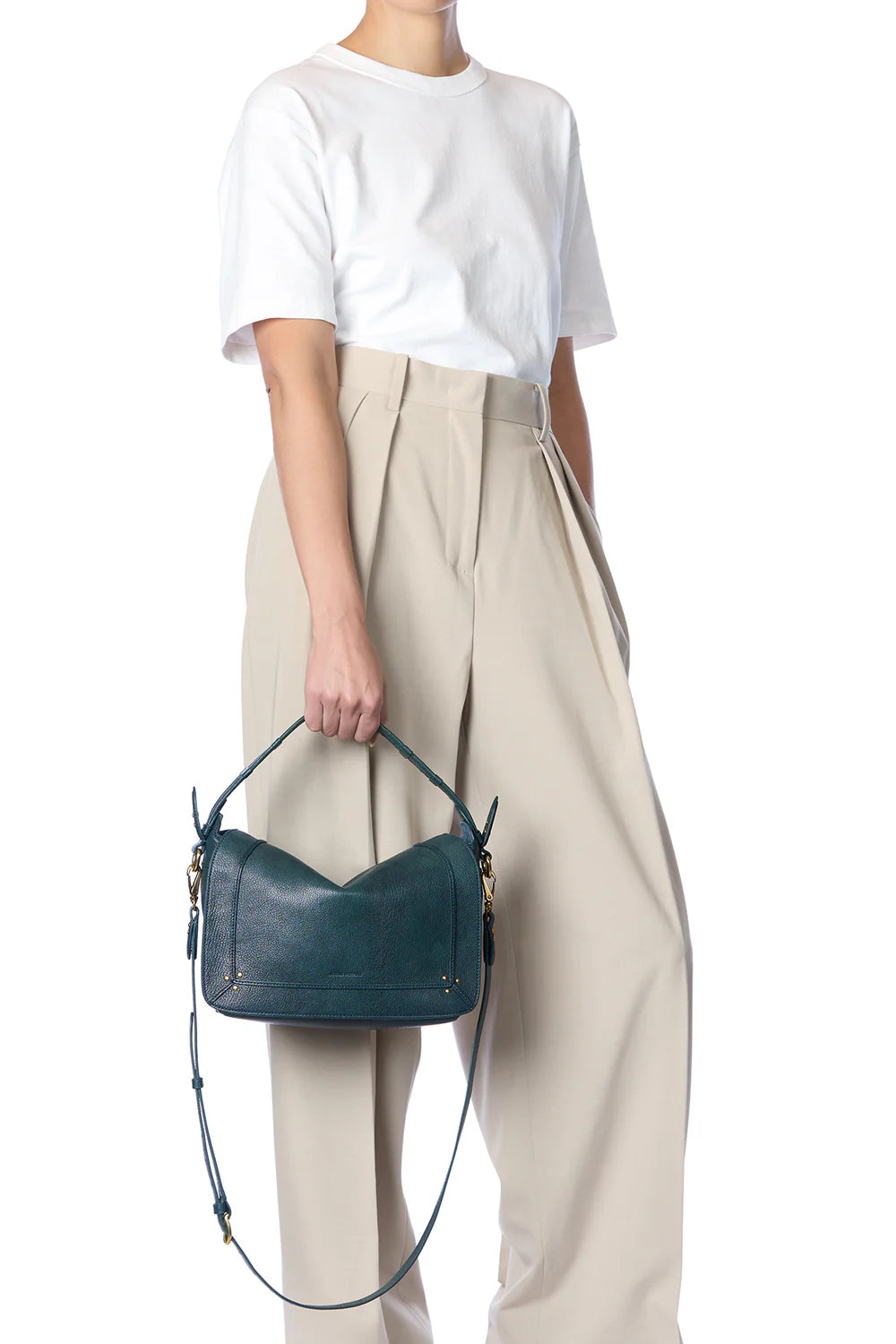 A person in a white t-shirt and high-waisted beige trousers holds the Jerome Dreyfuss Pepito Bag, a dark green leather handbag with a top handle and shoulder strap. The person’s face is not shown.