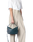 A person in a white t-shirt and high-waisted beige trousers holds the Jerome Dreyfuss Pepito Bag, a dark green leather handbag with a top handle and shoulder strap. The person’s face is not shown.